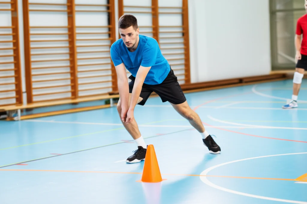 Atleta di pallavolo maschile durante un test di agilità T-test in palestra, catturato mentre tocca un cono. Obiettivo grandangolare 24mm per includere l'intero percorso del test, velocità dell'otturatore elevata, tracciamento del movimento, luce ambientale della palestra.