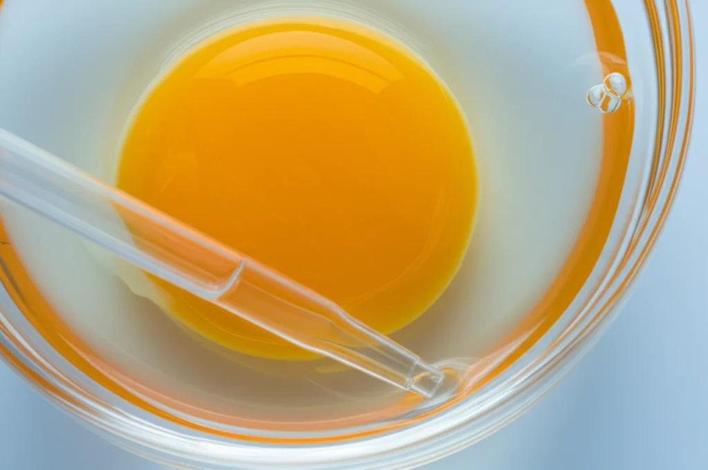 Interno di un uovo d'anatra rotto in una ciotola di vetro, con un albume denso e un tuorlo giallo intenso. Accanto, una pipetta che simboleggia l'analisi del lisozima. Illuminazione da laboratorio, precisa messa a fuoco sull'albume, obiettivo macro 100mm.