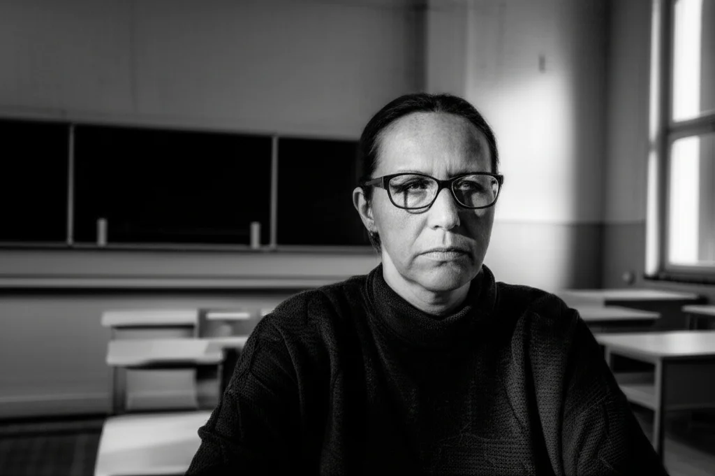 Ritratto di un'insegnante dall'aspetto preoccupato in un'aula vuota, obiettivo 50mm, profondità di campo ridotta che sfoca lo sfondo, illuminazione controllata che crea ombre drammatiche, bianco e nero filmico per enfatizzare l'emozione.