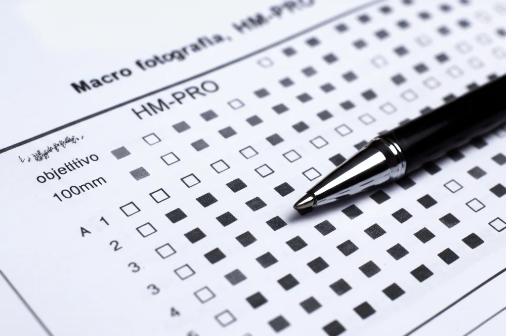 Macro fotografia, obiettivo 100mm, di un questionario HM-PRO cartaceo con una penna appoggiata sopra. La luce è controllata e laterale per evidenziare la texture della carta e le domande stampate. Messa a fuoco precisa su una sezione del questionario con le opzioni di risposta Likert. Alta definizione dei dettagli.