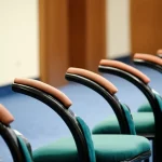 Immagine simbolica del dissenso istituzionale: una sedia vuota leggermente discostata da una fila di sedie identiche in una sala riunioni formale, luce direzionale che evidenzia la sedia vuota, obiettivo 50mm, profondità di campo ridotta.