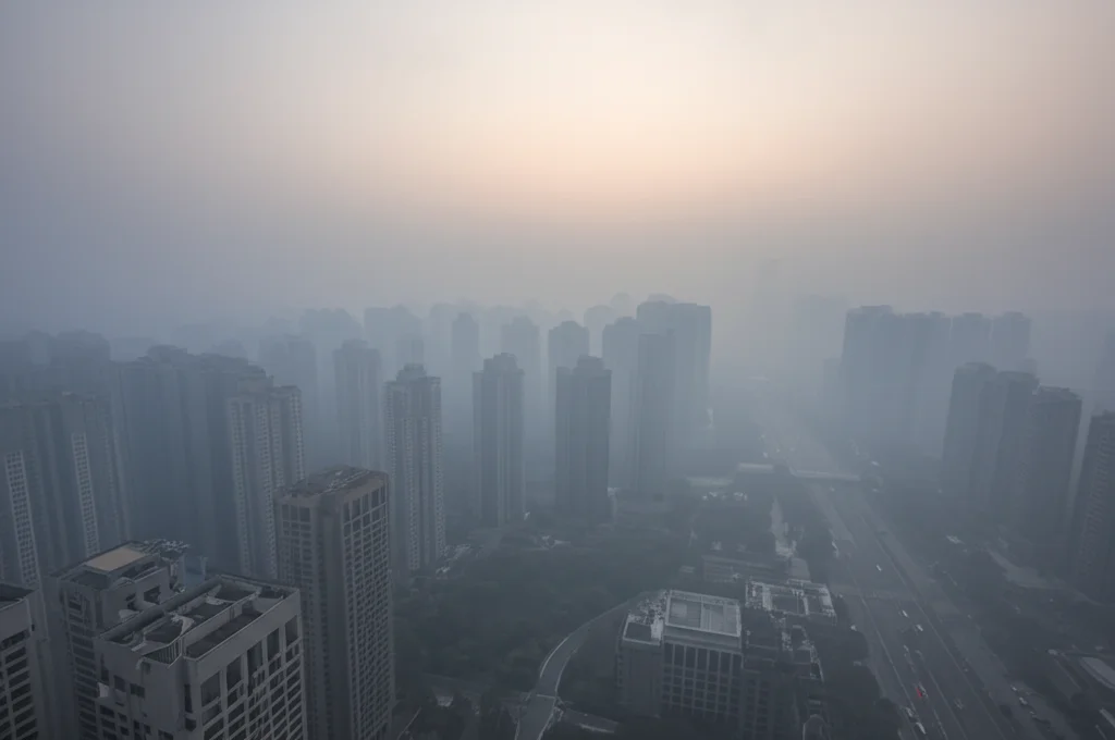 Veduta aerea di una metropoli cinese avvolta da una foschia densa di smog, simbolo dell'inquinamento da PM2.5. Obiettivo grandangolare 24mm, luce soffusa del tardo pomeriggio, focus nitido sugli edifici che emergono dalla foschia, long exposure per accentuare l'immobilità dell'inquinamento.