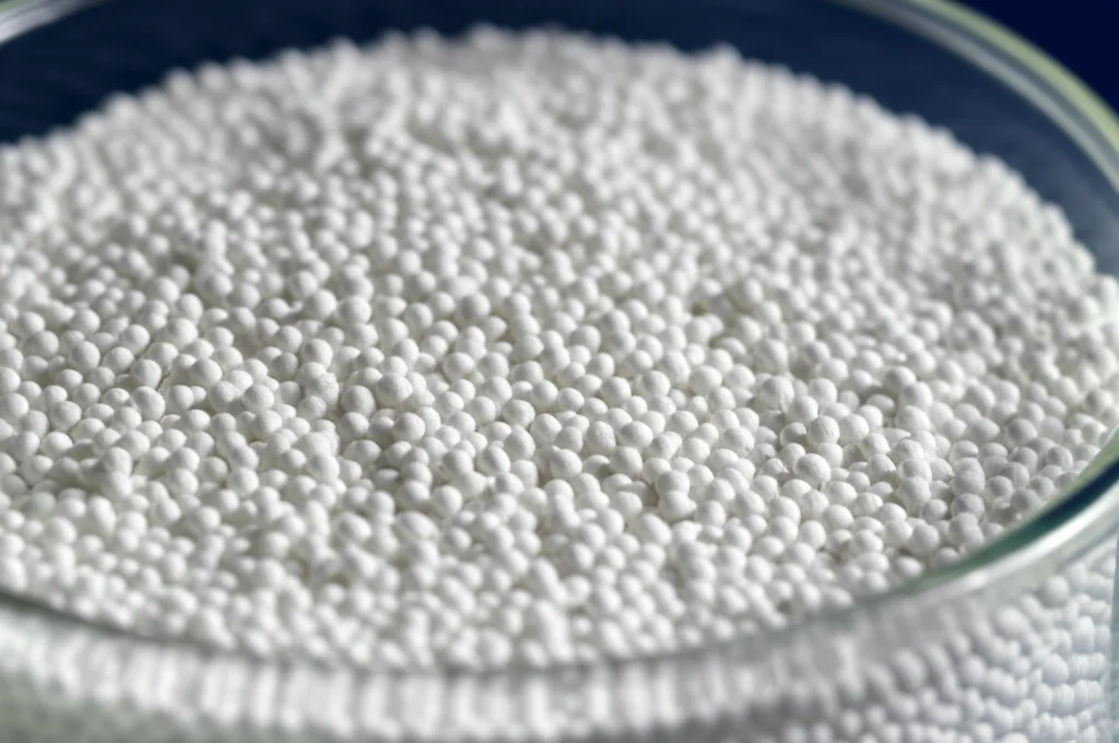 Primo piano macro, obiettivo 90mm, di setacci molecolari bianchi (sfere porose di 4 Angstrom) in un contenitore di vetro da laboratorio, con illuminazione laterale che ne evidenzia la texture porosa, simboleggiando l'assorbimento e la purificazione.