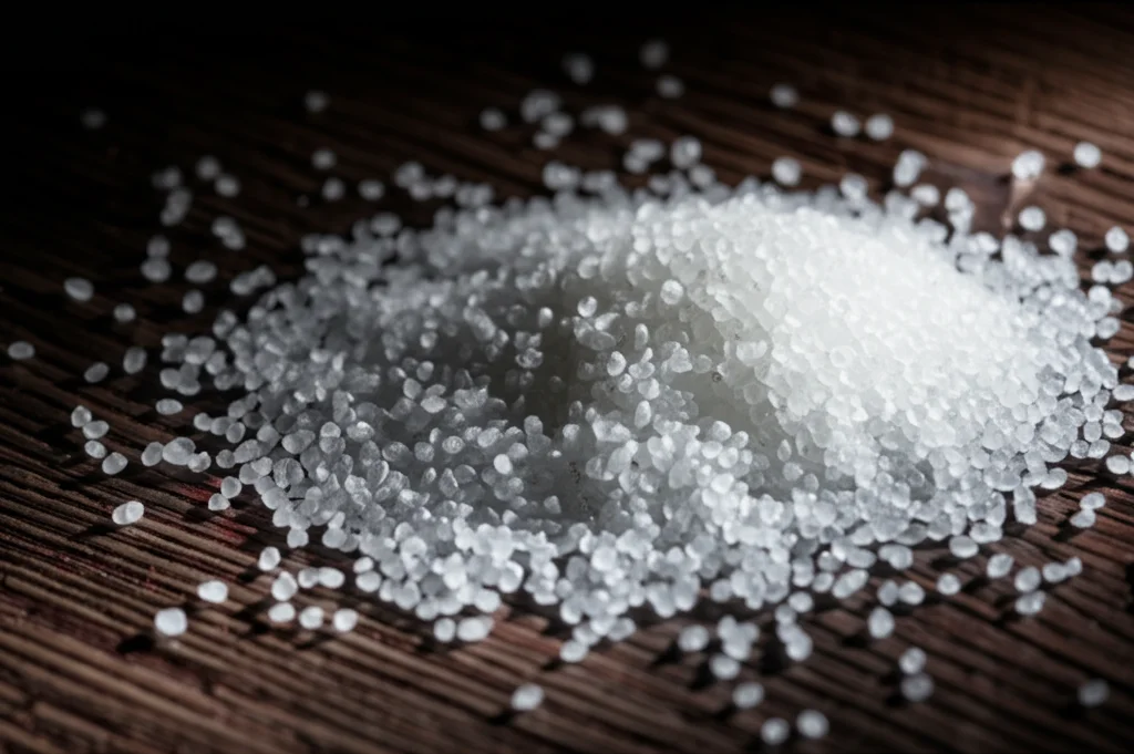 Fotografia still life, obiettivo macro 60mm, di piccoli granuli bianchi traslucidi di polimero biodegradabile (PLA modificato con TMC) accumulati su una superficie di legno scuro, alta definizione, illuminazione drammatica laterale per creare ombre e texture, simboleggiando il prodotto finale purificato e pronto all'uso.