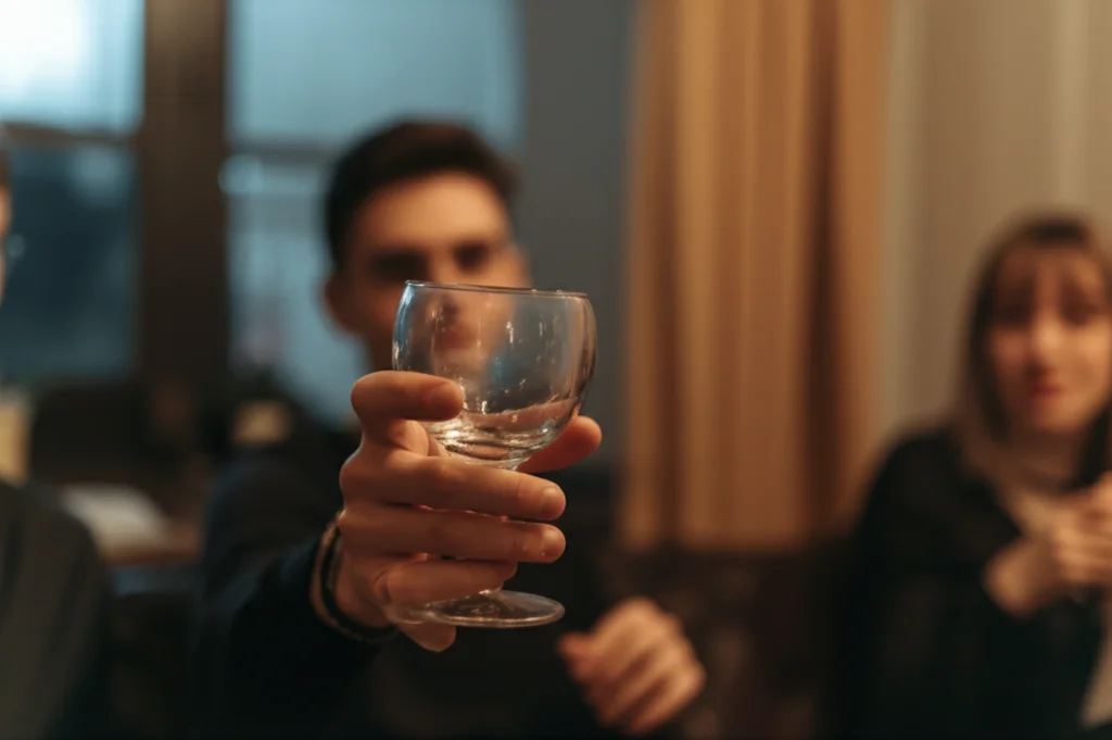 Scatto macro, obiettivo 100mm, di una mano adolescente che rifiuta discretamente un bicchiere di alcol offerto fuori campo durante una festa in casa, illuminazione controllata e calda. Alto dettaglio sulla texture della mano e del bicchiere.