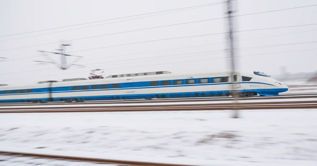 Fotografia paesaggistica di un treno ad alta velocità che sfreccia attraverso un paesaggio invernale cinese con neve leggera sui binari, obiettivo grandangolare 20mm, messa a fuoco nitida, leggero effetto mosso del treno per indicare velocità.
