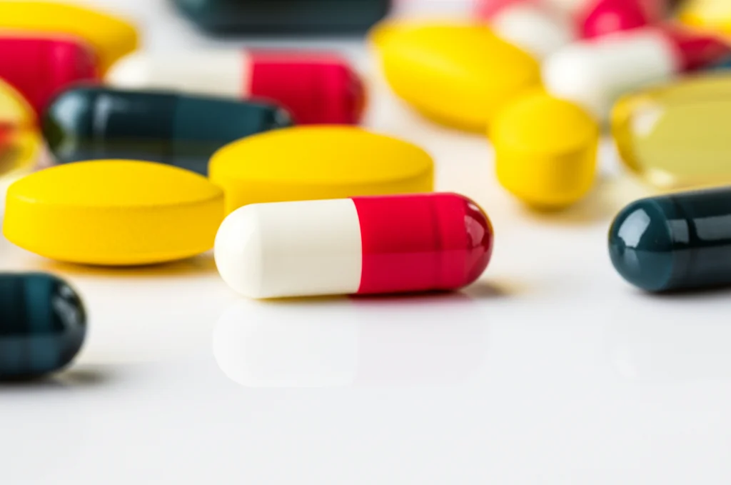 Primo piano di diverse pillole e capsule farmaceutiche colorate sparse su una superficie di laboratorio pulita e bianca, lente macro 70mm, alta definizione, illuminazione da studio controllata, focus preciso su una capsula rossa e bianca al centro.