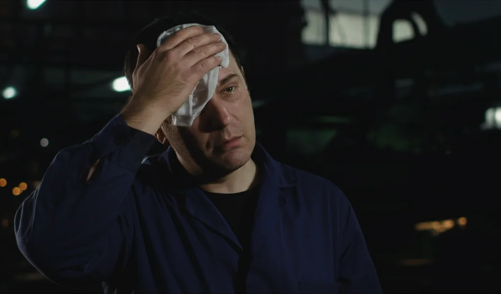 Uomo di mezza età, visibilmente stanco, che lavora di notte in una fabbrica poco illuminata, si asciuga il sudore dalla fronte. Obiettivo 35mm, stile film noir con forti contrasti tra luci e ombre, profondità di campo per mettere a fuoco l'uomo e sfocare leggermente lo sfondo industriale.