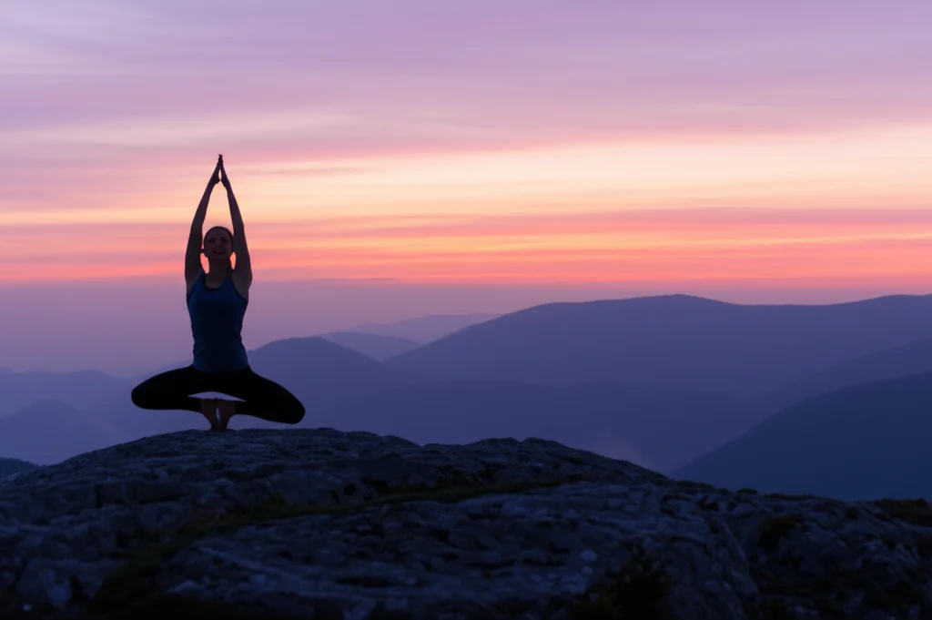 Fotografia sportiva di una persona che pratica yoga all'alba su una montagna, teleobiettivo zoom 200mm per comprimere la scena e mettere in risalto la figura contro il cielo colorato, velocità dell'otturatore moderata per catturare la posa stabile ma fluida, simboleggia equilibrio, consapevolezza (mindfulness) e connessione con il momento presente.