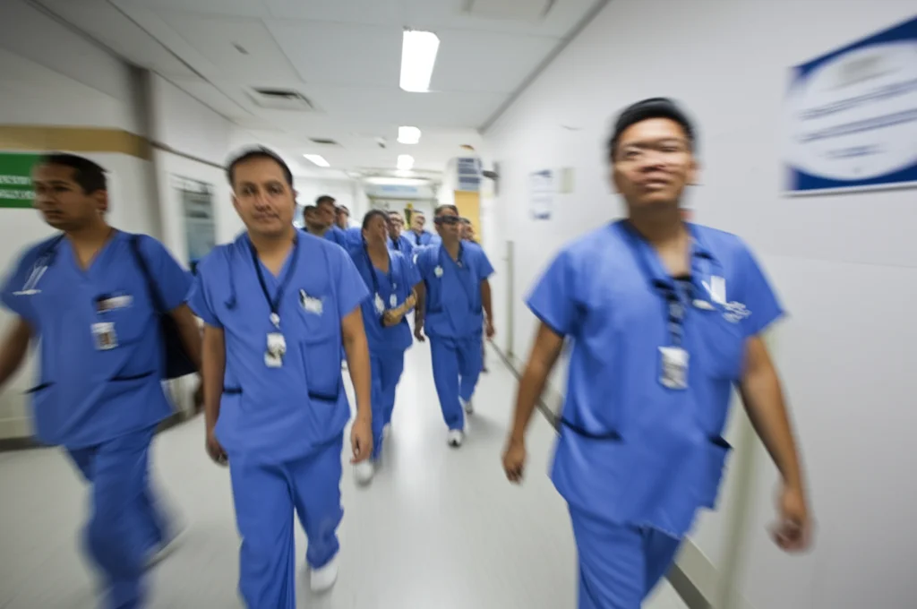 Foto dinamica di un gruppo di infermieri filippini che camminano frettolosamente in un corridoio d'ospedale affollato. Teleobiettivo zoom 100mm, velocità dell'otturatore elevata per congelare il movimento, tracciamento del movimento, luce fluorescente ospedaliera.