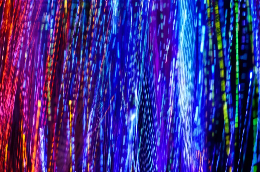 Visualizzazione di dati astratti rappresentanti i tre dataset (NSL-KDD, CICIDS-2017, RoEduNet-SIMARGL2021) come flussi digitali colorati che convergono verso un modello DNN, fotografia concettuale, obiettivo wide-angle 20mm, lunga esposizione per creare scie luminose.