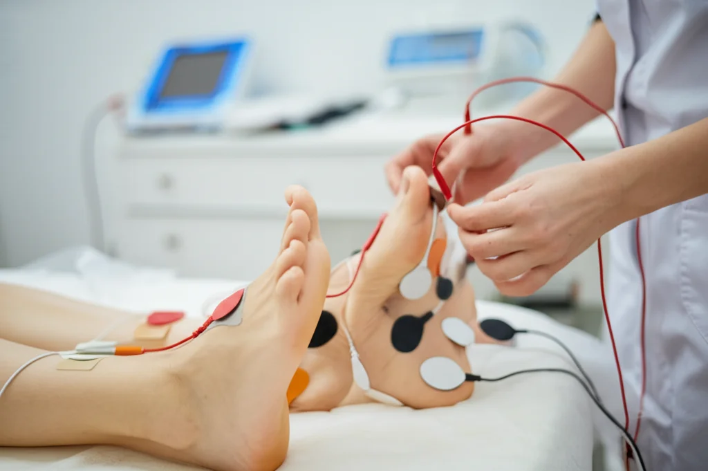 Analisi di impedenza bioelettrica (BIA) eseguita su una donna sdraiata su un lettino medico, elettrodi applicati sulla mano e sul piede destro, focus sui dettagli degli elettrodi con luce clinica controllata, obiettivo macro 100mm, alta definizione dei dettagli.