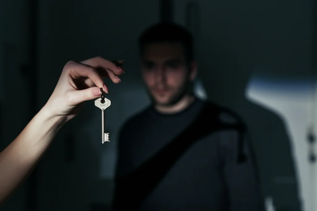 Immagine simbolica fotorealistica: una mano femminile che cerca di raggiungere una chiave appesa appena fuori dalla sua portata, mentre una figura maschile sfocata e in ombra osserva sullo sfondo. L'ambientazione è interna, scarsamente illuminata. Utilizzare un obiettivo macro 100mm per focalizzarsi sulla mano e la chiave, con alta definizione e illuminazione controllata per creare un'atmosfera tesa.