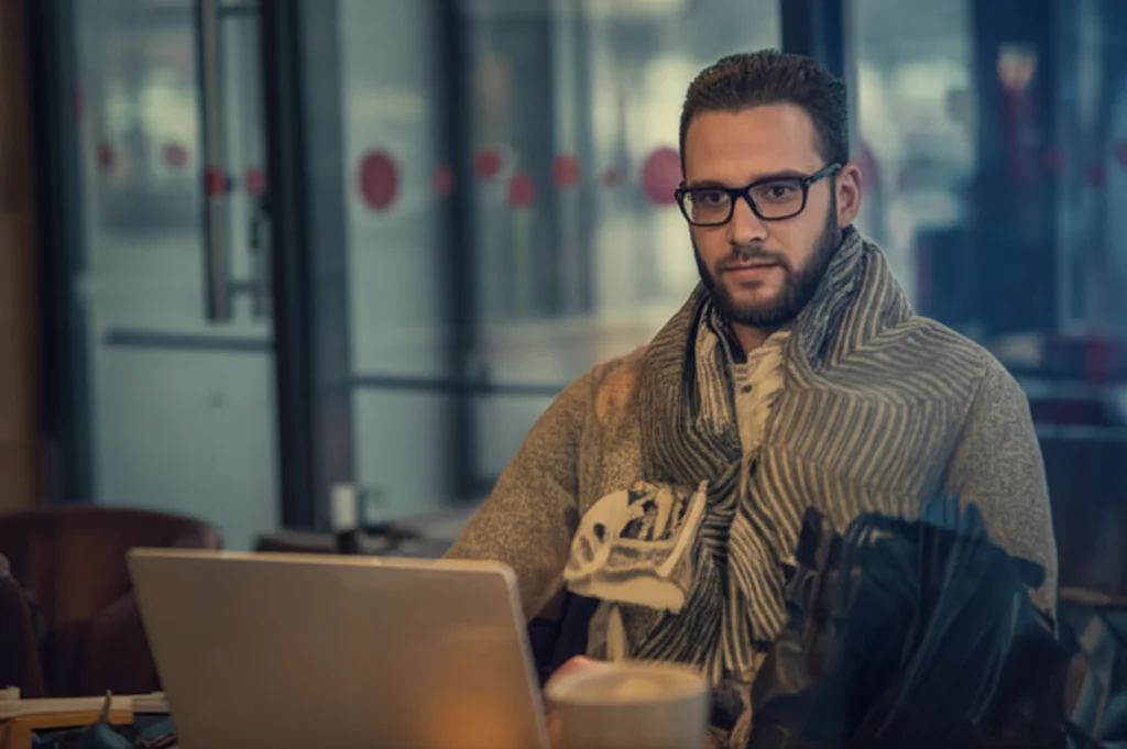 Fotografia di ritratto, obiettivo 50mm, di un giovane lavoratore della gig economy (es. grafico freelance) che lavora al computer portatile in un caffè affollato, sfondo urbano sfocato, luce naturale dalla finestra, duotone seppia e blu per un'atmosfera malinconica.