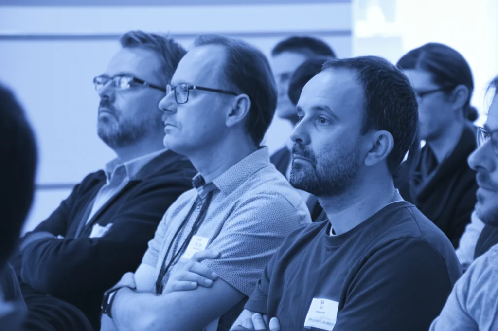 Fotografia di ritratto, obiettivo 35mm, che cattura un gruppo eterogeneo di cittadini attenti durante un incontro pubblico sulla politica energetica EGS. Illuminazione naturale e soffusa, profondità di campo focalizzata sulle espressioni di impegno riflessivo e leggera apprensione. Effetto duotone blu e grigio.