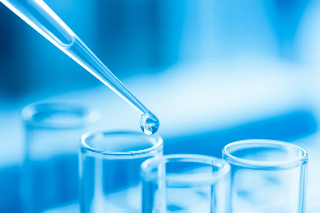 Immagine macro di una pipetta che rilascia un liquido bluastro in una provetta da laboratorio PCR, sfondo sfocato con attrezzatura scientifica moderna, obiettivo macro 100mm, illuminazione controllata da studio, messa a fuoco precisa sulla goccia e sulla punta della pipetta.