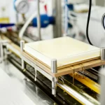 Primo piano di un sistema di microfiltrazione da laboratorio in funzione, tubi trasparenti con liquidi colorati (uno scuro tipo OMW, uno giallastro tipo CWE) che scorrono verso un modulo a membrana piatto visibile al centro, dettagli tecnici come manometri e valvole sullo sfondo sfocato, obiettivo prime 35mm, profondità di campo ridotta per focalizzare sulla membrana, illuminazione chiara da laboratorio.