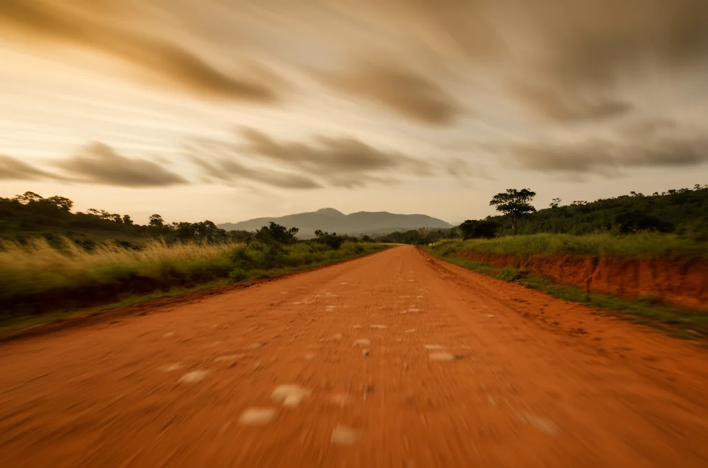 Fotografia grandangolare di una strada rurale sterrata nella Volta Region, Ghana, che si snoda verso colline lontane al tramonto. Obiettivo grandangolare 15mm, lunga esposizione per rendere il cielo leggermente mosso, messa a fuoco nitida sul paesaggio, colori caldi.