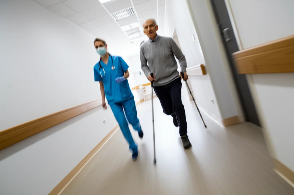 Scatto dinamico di un paziente anziano che esegue un test del cammino dei 6 minuti in un corridoio luminoso di ospedale, seguito da un operatore sanitario. Teleobiettivo zoom 100mm, velocità otturatore rapida per catturare il movimento, tracciamento del movimento.