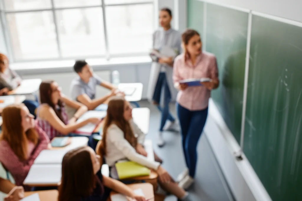 Fotografia di un'aula scolastica luminosa, vista leggermente dall'alto, studenti adolescenti diversi impegnati in una discussione con un insegnante sorridente vicino alla lavagna. Obiettivo prime 35mm, profondità di campo ridotta per sfocare leggermente lo sfondo, luce naturale dalle finestre.