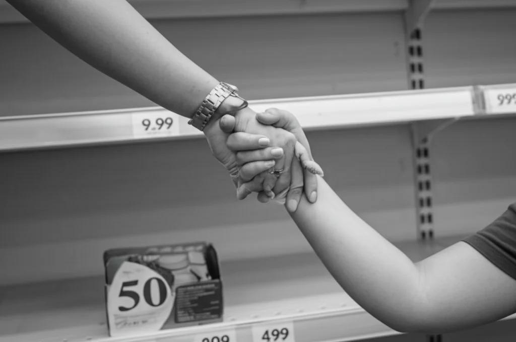 Una mano adulta tiene stretta una mano di bambino davanti a uno scaffale di supermercato quasi vuoto, obiettivo prime 50mm, profondità di campo, luce soffusa, bianco e nero film.