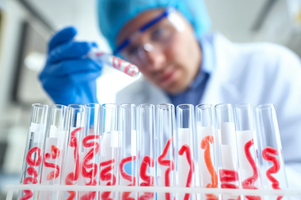 Fotografia di laboratorio che mostra diverse provette contenenti campioni di tessuto intestinale di topo da modelli di cancro del colon-retto, con un ricercatore in background sfocato. Obiettivo prime 35mm, profondità di campo ridotta.