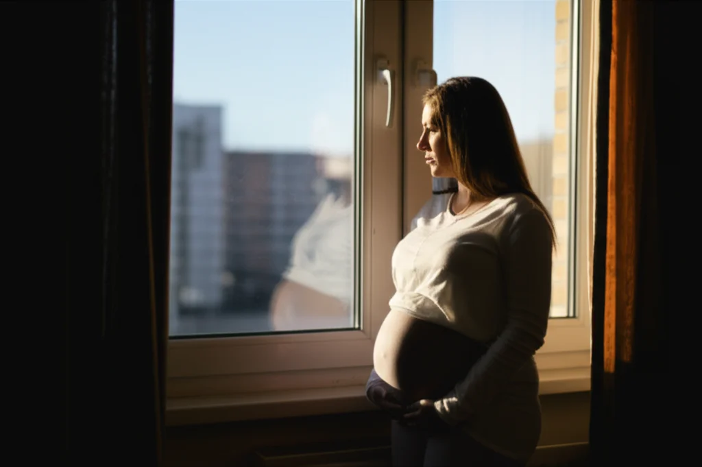 Ritratto fotografico di una donna incinta che guarda pensierosa fuori dalla finestra durante il lockdown, luce naturale soffusa che entra dalla finestra, stile intimo e riflessivo, obiettivo 35mm, profondità di campo che sfoca leggermente lo sfondo esterno.