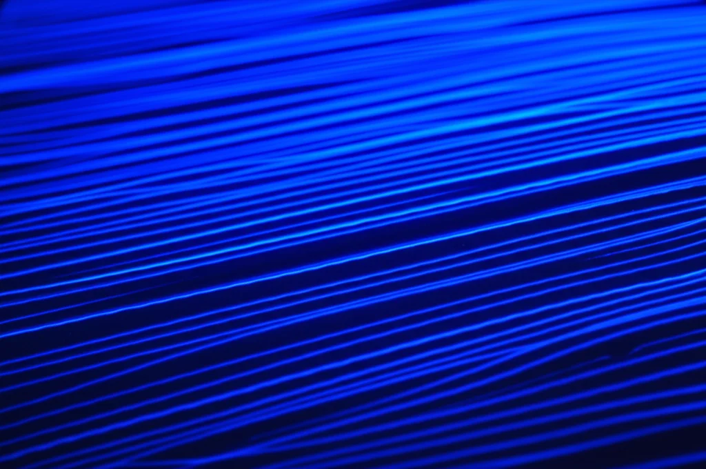 Visualizzazione scientifica astratta del flusso d'aria turbolento su una superficie piana, con micro-onde longitudinali bluastre che viaggiano lungo la superficie, ispirate alla pelle di delfino. Illuminazione controllata per evidenziare le micro-strutture e le onde, obiettivo macro 100mm, alta definizione, messa a fuoco precisa.