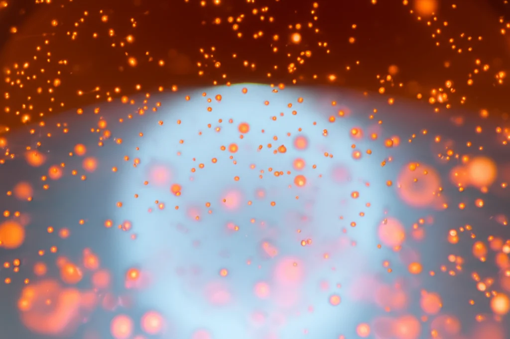 Macro fotografia, 85mm lens, di nanoparticelle rosso-arancio luminescenti sospese in un liquido trasparente all'interno di un becher da laboratorio, messa a fuoco precisa sulle particelle, illuminazione controllata da dietro che ne evidenzia la dispersione, alto dettaglio della texture liquida.