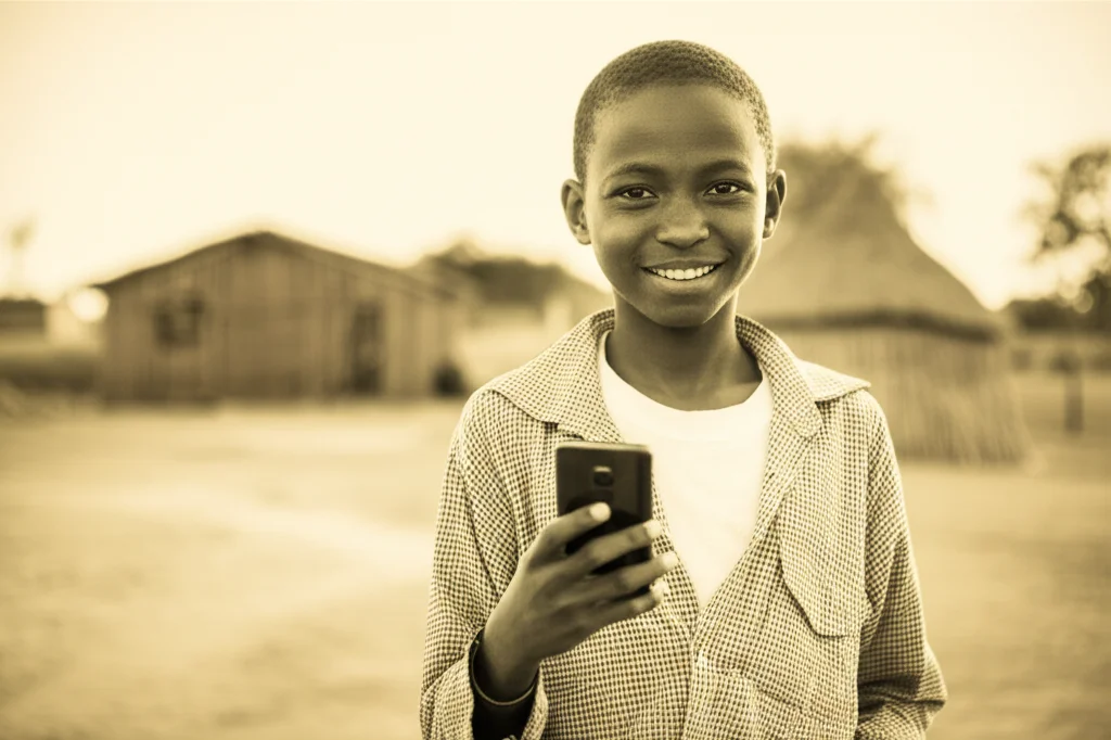 Fotografia ritratto di un adolescente keniota sorridente che tiene in mano uno smartphone, luce naturale del tardo pomeriggio, obiettivo 35mm, profondità di campo ridotta per sfocare lo sfondo di un villaggio, duotone caldo seppia e ocra.