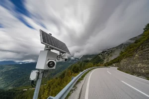 Fotografia grandangolare di una strada di montagna con un sistema di rilevamento frane (telecamera, pannello solare) installato sul lato, messa a fuoco nitida, lunga esposizione per nuvole setose, obiettivo grandangolare 10mm, che illustra l'applicazione pratica della tecnologia YOLOv8-GCB.