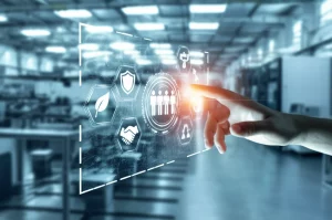 Immagine fotorealistica che simboleggia l'Industria 5.0: una mano umana che sfiora delicatamente un'interfaccia digitale olografica fluttuante nello spazio. L'interfaccia mostra icone luminose interconnesse rappresentanti la sostenibilità (una foglia stilizzata), la resilienza (uno scudo protettivo) e la collaborazione uomo-macchina (sagome stilizzate di una persona e un robot che si danno la mano). Sullo sfondo, un ambiente industriale moderno, pulito e luminoso, leggermente sfocato. Obiettivo prime 35mm, profondità di campo ridotta per focalizzare sull'interazione mano-ologramma, tonalità blu e verde duotone che evocano tecnologia e natura.
