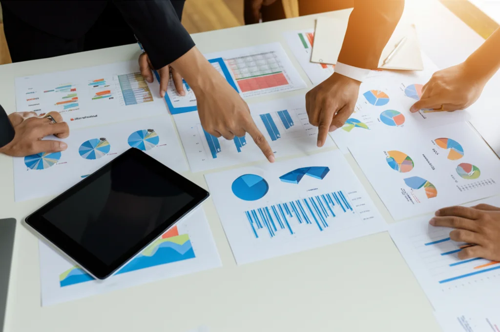 Fotografia dall'alto di un tavolo da riunione con grafici, matrici di valutazione stampate (simili a AHP/TOPSIS) e mani di persone che indicano i dati. Luce naturale da finestra laterale, obiettivo 24mm grandangolare moderato, focus nitido sui documenti.