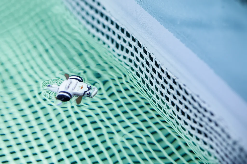 Fotografia macro, obiettivo 90mm, di un singolo pesce robotico che nuota vicino a una parete porosa a rete in un'arena circolare d'acqua. Illuminazione controllata che evidenzia la texture della rete e le piccole onde sulla superficie dell'acqua. Messa a fuoco precisa sul pesce e sull'angolo che forma con la parete.