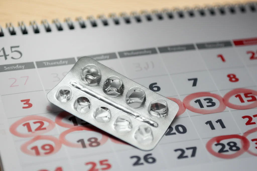 Primo piano di un calendario da tavolo con alcuni giorni cerchiati, accanto a un blister quasi vuoto di pillole contraccettive, focus selettivo sul calendario, luce naturale morbida da una finestra, lente macro 60mm, alta definizione.