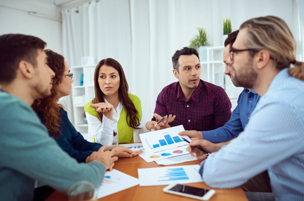 Fotografia realistica di un gruppo eterogeneo di adulti (genitori e non) seduti attorno a un tavolo, che discutono animatamente ma costruttivamente guardando dei grafici. Obiettivo 35mm, luce ambientale morbida, profondità di campo media per includere diverse espressioni facciali.