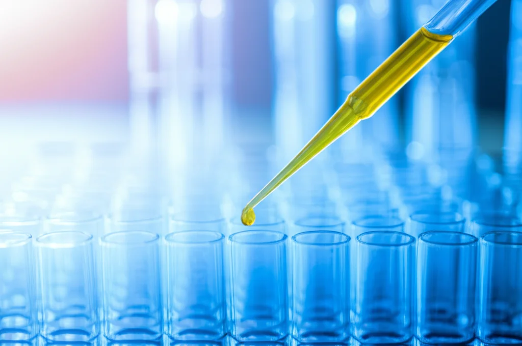 Primo piano macro di una fiala di campione di DNA in un laboratorio di genetica, messa a fuoco precisa sulla punta della pipetta che aggiunge un reagente, illuminazione controllata da laboratorio, obiettivo macro 100mm, sfondo leggermente sfocato con altre attrezzature scientifiche.