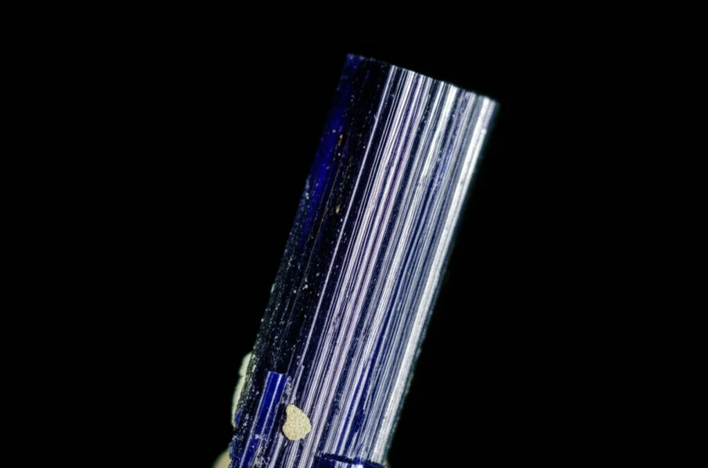 Macro fotografia di un cristallo di riebeckite blu scuro, allungato e prismatico, illuminato lateralmente per evidenziare le facce cristalline e la lucentezza. Obiettivo macro 90mm, alta definizione, sfondo nero sfocato.