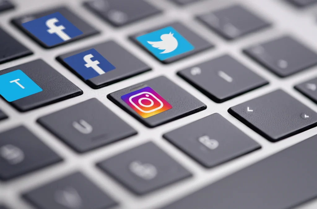 Fotografia macro di una tastiera di laptop con piccole icone di social media (Facebook, Instagram, Twitter) sovrapposte leggermente sfocate sullo sfondo. Obiettivo macro 90mm, messa a fuoco precisa sui tasti 'S' 'M' 'E', illuminazione controllata laterale che crea ombre morbide, dettaglio elevato sulla texture dei tasti.