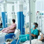 Immagine fotorealistica di un medico o farmacista in ambiente ospedaliero che regola con attenzione una flebo di Remdesivir collegata a un paziente COVID-19. Sullo sfondo, altri pazienti diversi per età ed etnia ricevono cure, suggerendo personalizzazione. Utilizzo di un obiettivo prime (24-35mm) con profondità di campo (depth of field) per mettere a fuoco l'azione sulla flebo ma mantenere visibile il contesto. Illuminazione controllata, stile clinico ma umano.