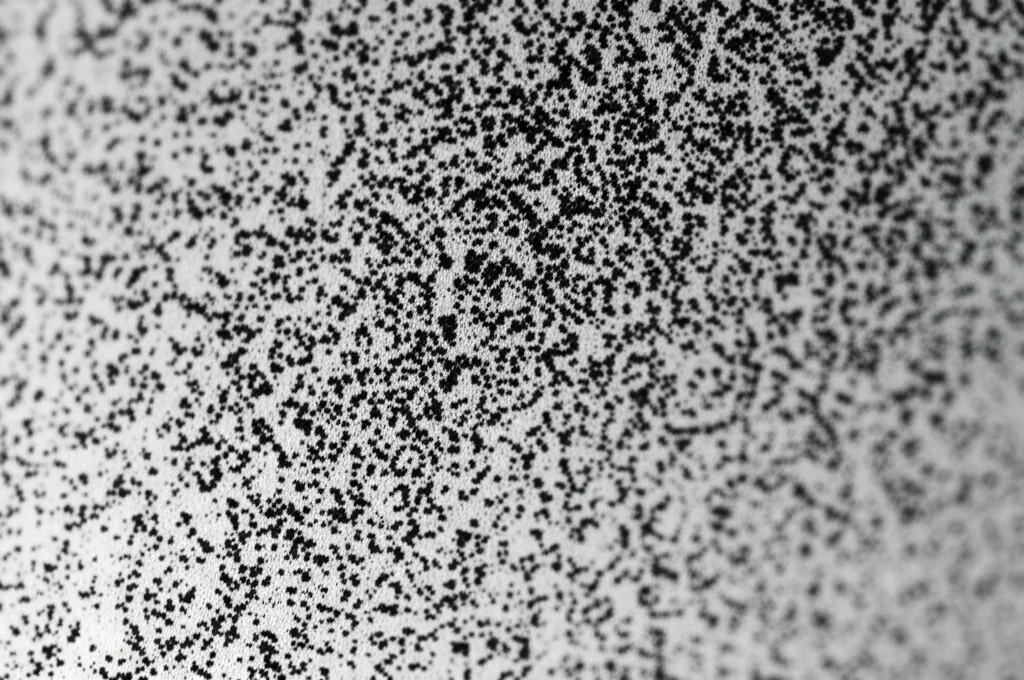 Macro fotografia di particelle scure simili a fuliggine depositate su una superficie metallica chiara e leggermente riflettente, obiettivo macro 100mm, alta definizione, illuminazione controllata laterale per evidenziare la texture granulare delle particelle.