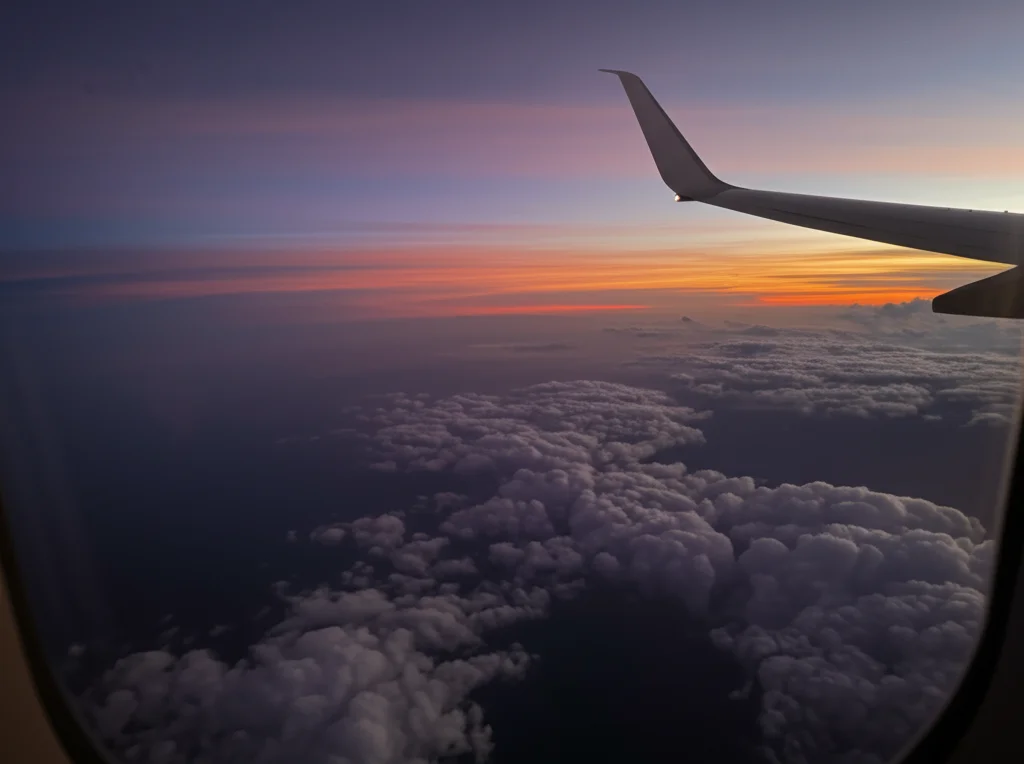 Vista dal finestrino di un aereo che sorvola le nuvole al tramonto, colori caldi e morbidi, obiettivo 50mm, messa a fuoco sulle nuvole lontane, evocando il tema del viaggio di ritorno e della riflessione dopo un'esperienza difficile.