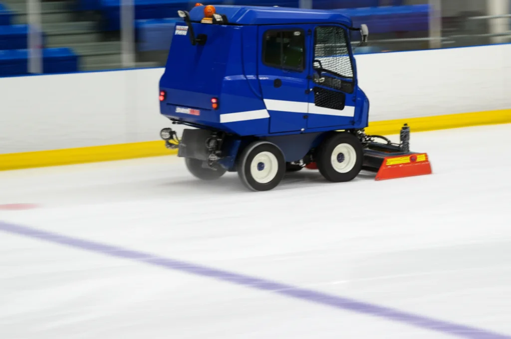 Fotografia d'azione di una macchina resurfacing (tipo Zamboni) vista di tre quarti mentre lavora su una pista di hockey vuota, getto d'acqua visibile sul retro, teleobiettivo 200mm, tracciamento del movimento, velocità otturatore elevata per congelare l'azione ma con leggero motion blur delle ruote e della lama posteriore.