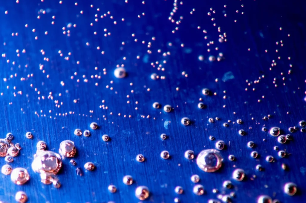 Primo piano macro di una superficie metallica in acciaio X70 durante il processo di elettrodeposizione in una cella elettrolitica. Si vedono bollicine salire dalla superficie immersa in una soluzione bluastra (solfato di rame e stagno). Illuminazione controllata da laboratorio, obiettivo macro 100mm, alta definizione dei dettagli sulla superficie del metallo e nella soluzione, messa a fuoco precisa.