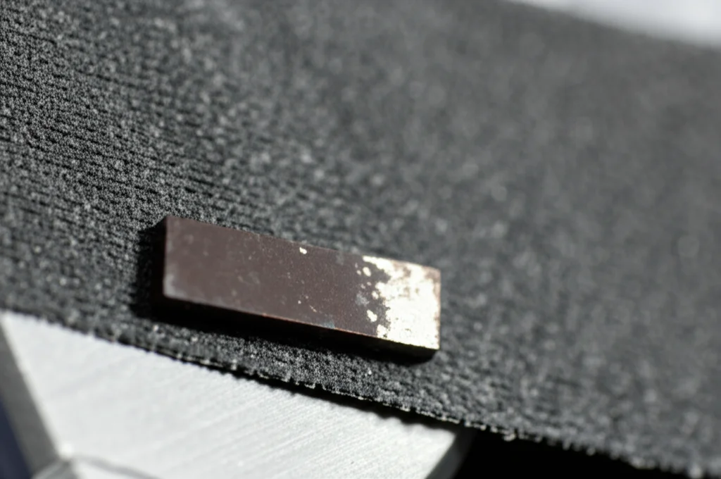 Immagine ravvicinata di un test di abrasione con carta vetrata su un campione di acciaio rivestito con la lega Cu-Sn. Si vede il campione metallico scuro premuto contro la carta vetrata grigia (grana 2000). Dettagli sulla texture della carta e sulla superficie del rivestimento che mostra una leggera usura ma mantiene la sua integrità. Obiettivo macro 60mm, illuminazione da laboratorio focalizzata sull'area di contatto, alta definizione.