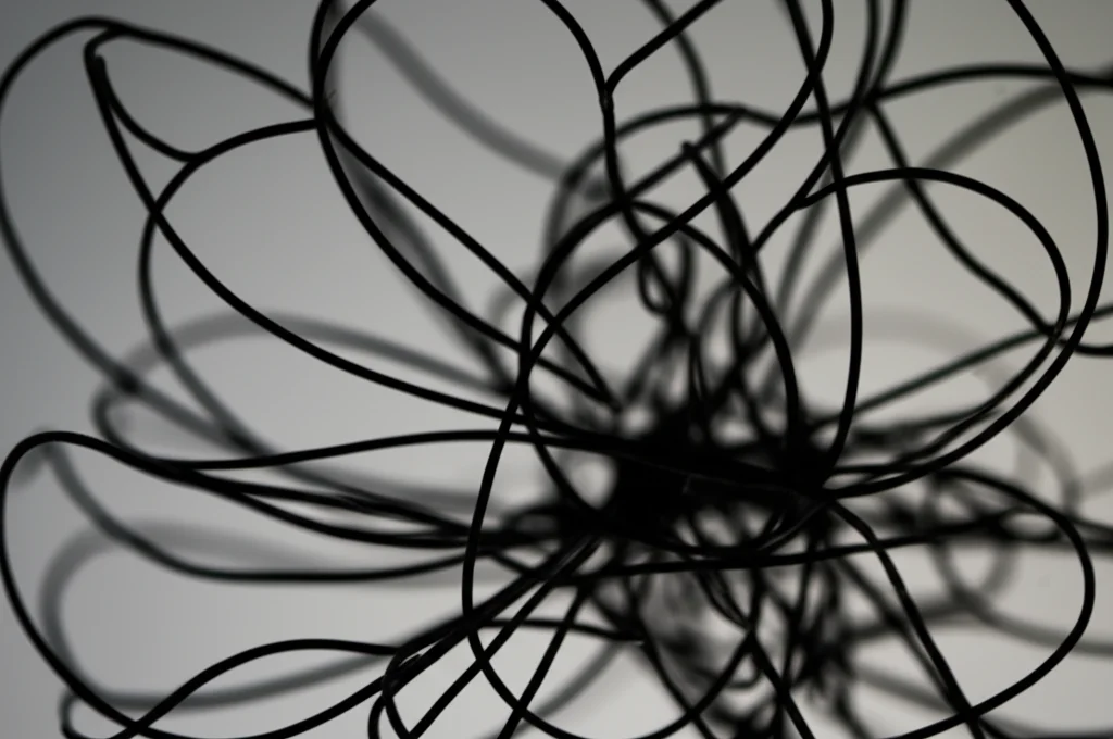 Fotografia macro (obiettivo macro 100mm) di un groviglio di fili aggrovigliati e scuri su uno sfondo neutro e sfocato. Illuminazione controllata per creare forti contrasti e dettagli elevati sui fili, simboleggiando confusione e stress mentale.