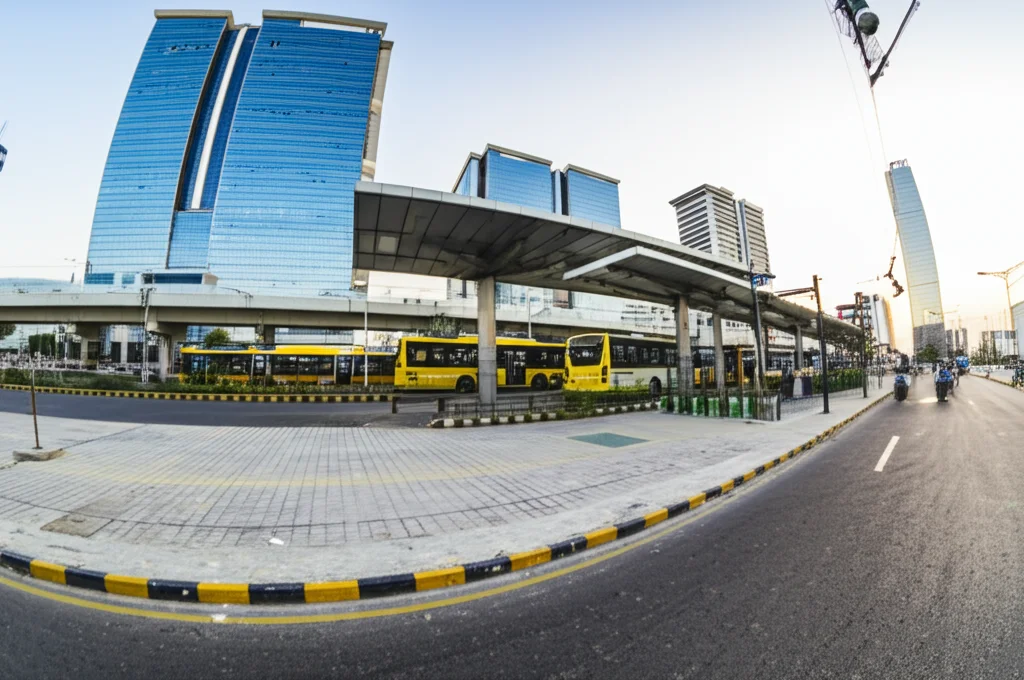 Fotografia grandangolare di un moderno hub di trasporto intermodale in una città pakistana, con autobus a basse emissioni, una stazione di bike sharing e forse un treno leggero in lontananza. Obiettivo grandangolare 18mm, messa a fuoco nitida, luce del tardo pomeriggio per un'atmosfera dinamica.