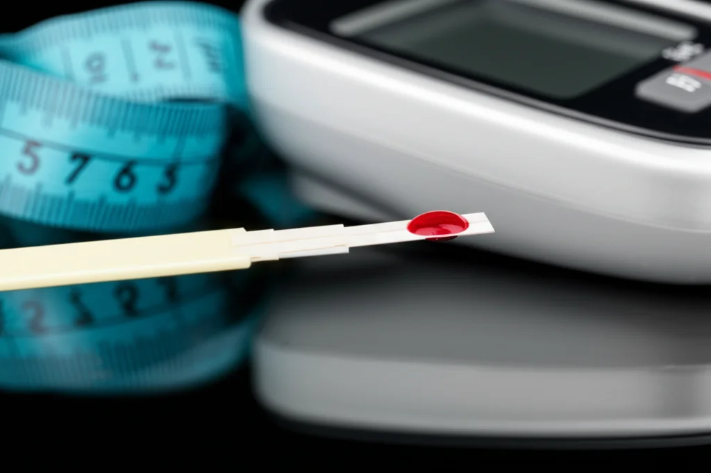 Fotografia macro di una goccia di sangue su una striscia reattiva per la glicemia, con un misuratore di pressione e un centimetro da sarta sullo sfondo sfocato. Obiettivo macro 100mm, alta definizione, illuminazione controllata da studio.