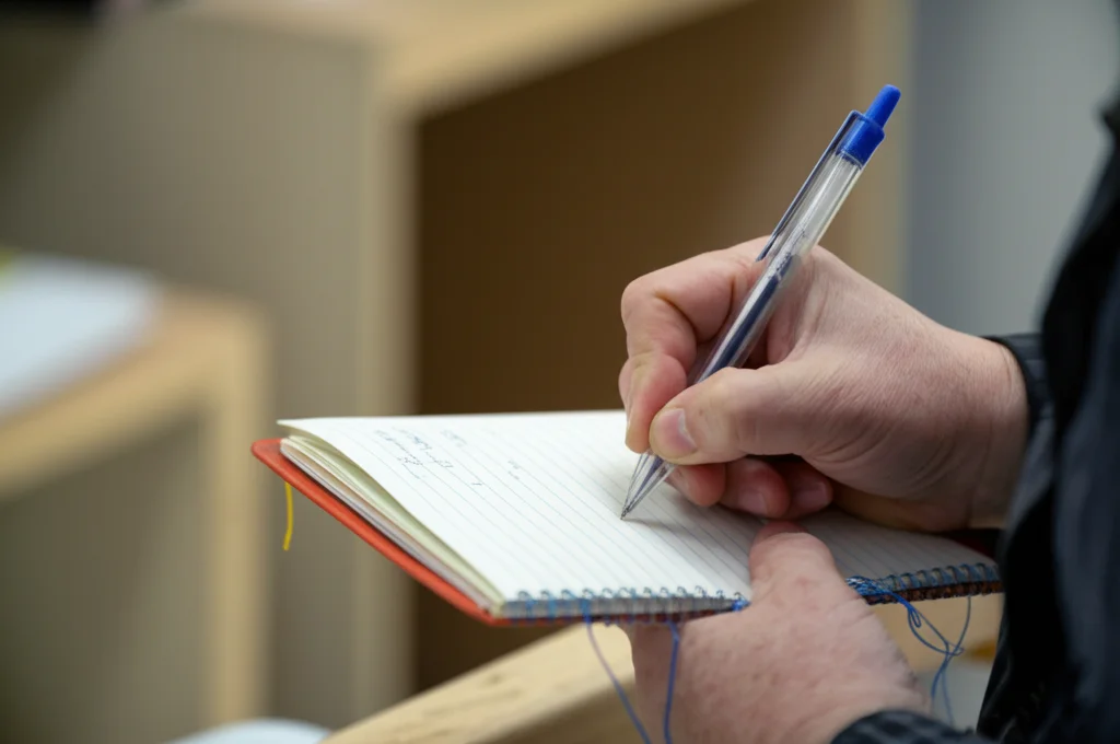 Dettaglio macro di una mano che scrive lentamente appunti su un taccuino consumato, con sfondo sfocato di un ambiente di ricerca sul campo. Obiettivo macro 100mm, messa a fuoco precisa sulla mano e sul taccuino, luce ambientale controllata, alta definizione.