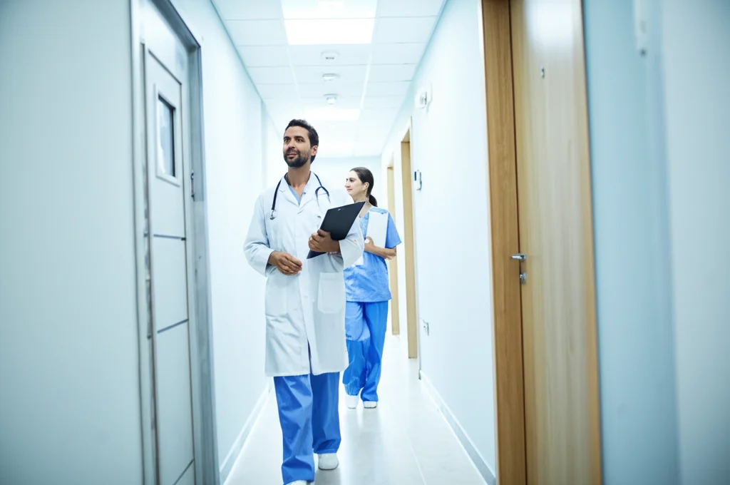 Fotografia grandangolare, un moderno e luminoso corridoio di uno studio medico con personale sanitario che cammina con passo deciso, simboleggiando progresso e accessibilità alle cure. Obiettivo wide-angle 15mm, messa a fuoco nitida, luce brillante.